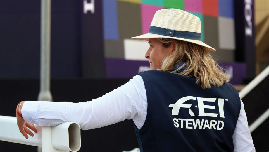 An FEI steward leans on white railings at a horse show.