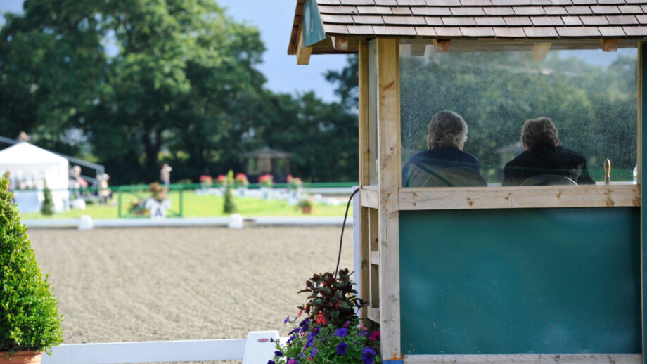 A judges’ box pictured from behind with a judge and their writer sitting inside facing away from the camera, with an outdoor dressage arena behind.