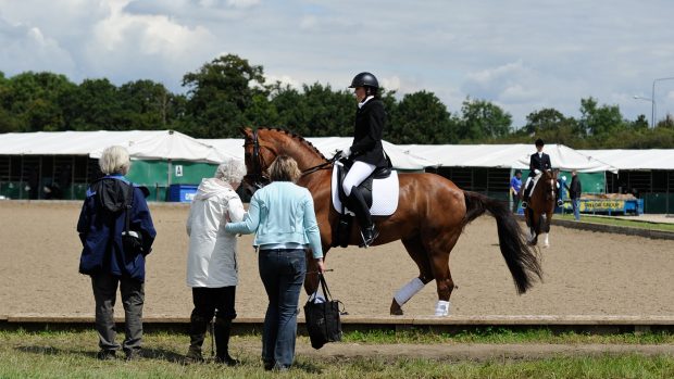 A rider in the foreground warming up a chestnut horse for a dressage show with three onlookers with their backs to camera looking on and stables in the background