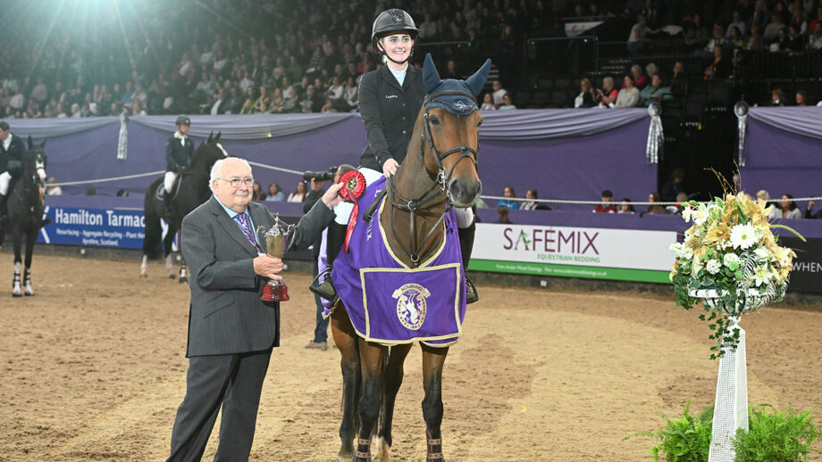 Jodie Hall McAteer being presented with a trophy and rosette on board her horse wearing a purple winners rug at HOYS 2024.